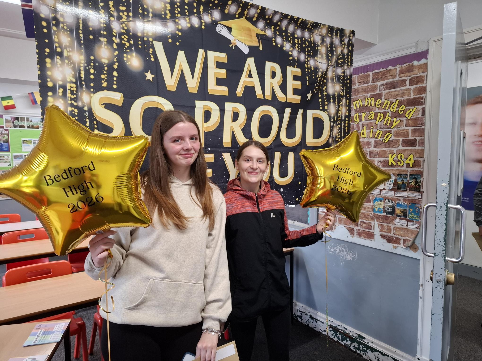 Two young women from Bedford High School with their results and in front of a banner, saying, 'we are so proud of you!'